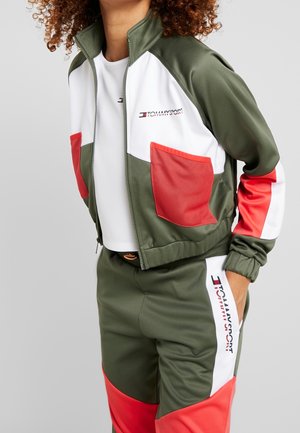 Femme portant une veste de survêtement verte, blanche et rouge avec des poches en filet et un pantalon assorti avec le logo Tommy Sport sur la veste et le pantalon.