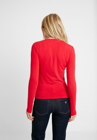 Camisa roja de manga larga hecha de tela suave, con un sencillo escote de cuello redondo. Llevada con jeans de mezclilla oscuros. Textura suave, diseño ajustado.