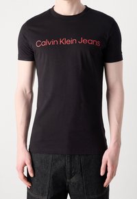 Černé bavlněné tričko s červeným logem "Calvin Klein Jeans" na hrudi. Krátké rukávy a kulatý výstřih s úzkým střihem.