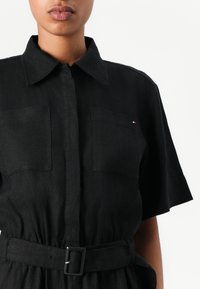 Vestido de camisa de lino negro con cuello de solapa, mangas cortas, dos bolsillos en el pecho y una cintura ceñida con cinturón. Presenta un pequeño detalle de logo.