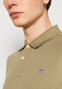 Chaki polo marškiniai su tekstūruota medžiaga, trimis mygtukais ir mažu išsiuvinėtu logotipu ant krūtinės.