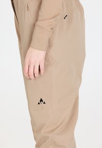 Giacca beige in tessuto con una vestibilità slim, dotata di una tasca con zip e un accento di logo nero vicino al fondo. Texture liscia e design su misura.