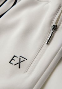 Biała bluza z materiału, z czarnym logo "EX" i srebrnym zamkiem błyskawicznym na kieszeni, prezentująca gładką fakturę i proste linie.