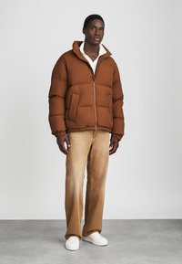 Giacca a vento marrone con chiusura zip, polsini elasticizzati, due tasche anteriori, indossata sopra una camicia crema, abbinata a pantaloni beige a gamba larga e sneakers bianche.