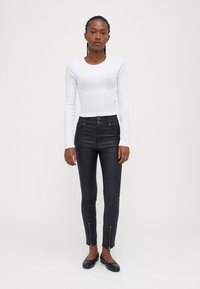 Witte langmouwtop en zwarte aangesloten jeans met ritsen bij de enkels. De jeans hebben een gladde textuur en een sluiting met twee knopen.