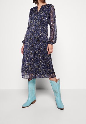 Mujer con un vestido midi azul estampado con mangas largas transparentes y botas de cowboy azul claro, de pie frente a un fondo blanco.