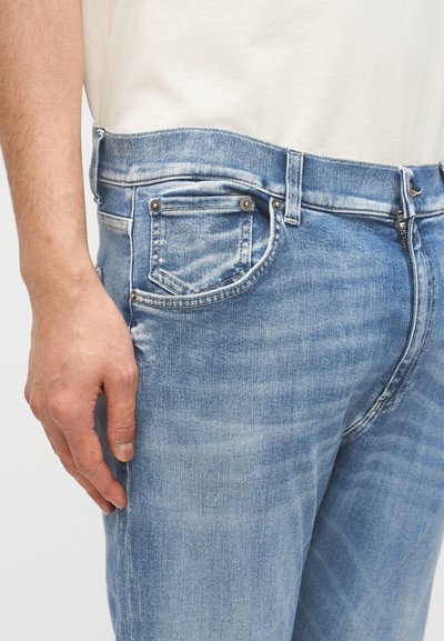 Κόντινο πλάνο ενός ατόμου που φοράει ανοιχτόχρωμα τζιν παντελόνια και λευκό πουκάμισο, με το ένα χέρι να ακουμπάει δίπλα στην τσέπη.