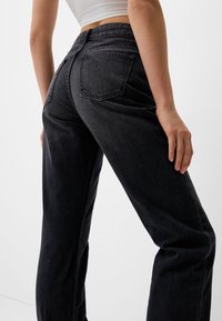 Jeans negros de tiro alto con perneras rectas, bolsillos traseros y una textura sutil. La tela parece duradera y ligeramente desgastada.