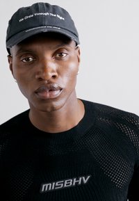 Pull noir en tricot avec un motif texturé et un logo "MISBHV." Casquette noire avec le texte blanc "We Circle Through The Night."