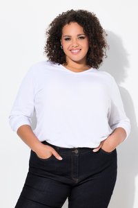 Femme souriante aux cheveux bouclés portant un chemisier blanc à manches longues et un jean foncé, debout les mains dans les poches devant un mur blanc.