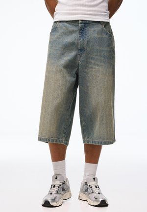 Személy fakó, laza, farmerszövetből készült térd hosszúságú rövidnadrágot, fehér sportzoknit és ezüstszürke atlétikai cipőt visel, fehér háttér előtt áll.