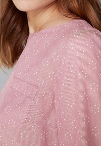 Blus av randig rosa tyg med ett subtilt mönster, som har små vita prickar, rund halsringning och en bröstficka.
