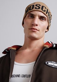 Ruskea huppari, jossa on raidallinen kaulus, musta ja oranssi Moschinon pipo sekä valkoinen logolla varustettu tanktop. Sujuva, rento kangas ja löysä istuvuus.