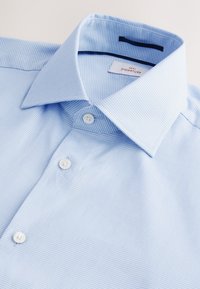 Chemise texturée bleu clair avec col pointu et boutons blancs, étalée à plat sur une surface blanche.