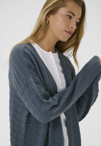 Femme aux longs cheveux blonds portant des boucles d'oreilles en argent, une chemise blanche à boutons et un cardigan en tricot texturé bleu, regardant pensivement vers le bas.