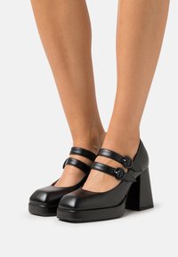 Black leather platform heels with two adjustable straps and round buttons, featuring a chunky block heel and rounded toe design.