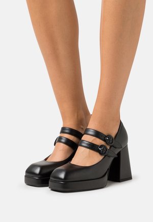 Escarpins à plateforme en cuir noir avec deux brides réglables et des boutons ronds, présentant un talon épais et un design de bout arrondi.