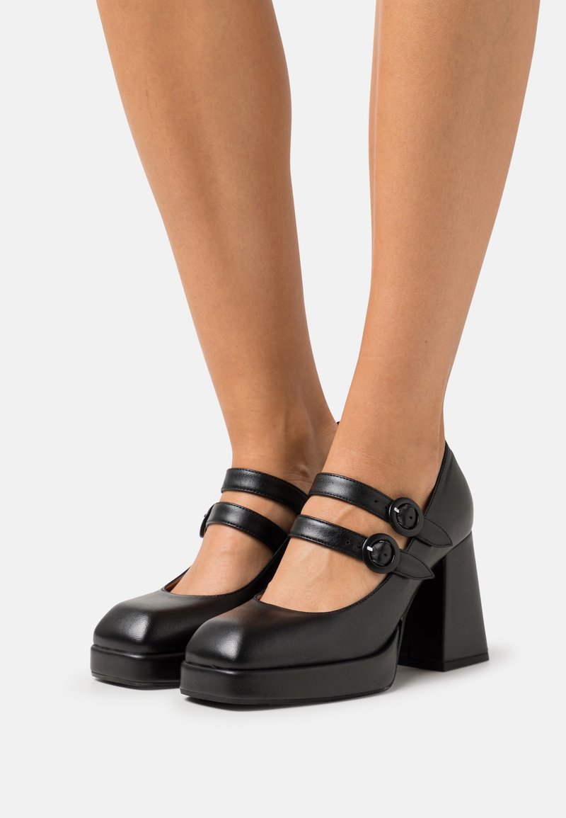 Black leather platform heels with two adjustable straps and round buttons, featuring a chunky block heel and rounded toe design.