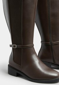Brązowe skórzane buty za kolano z okrągłym noskiem, kontrastującymi teksturowymi panelami, detalem w postaci paska na kostce oraz zamkiem błyskawicznym z boku.