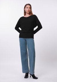 Juodas ribotu audiniu megztinis su plačia iškirpte, derinamas su šviesiai mėlynomis, laisvai krentančiomis džinsinėmis kelnėmis. Modelis dėvi juodus kulniukais apkurtus aulinukus.