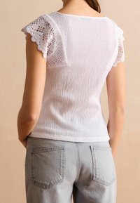 Haut blanc texturé avec des manches courtes en dentelle, associé à un jean gris clair avec deux poches arrière.