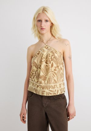 Donna bionda che indossa un top beige a collo halter con motivi marroni a forma di foglia e pantaloni marrone scuro, in piedi davanti a uno sfondo chiaro uniforme.