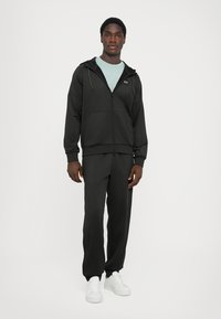 Svart zip-up hoodie med framfickor, dragsko på huven, kombinerad med matchande svarta joggers och vita sneakers. Modellen står i en neutral miljö.