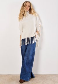 Poncho in maglia beige con colletto alto e dettagli in frange all'orlo, abbinato a jeans in denim blu a gamba larga e stivali alla caviglia marroni.