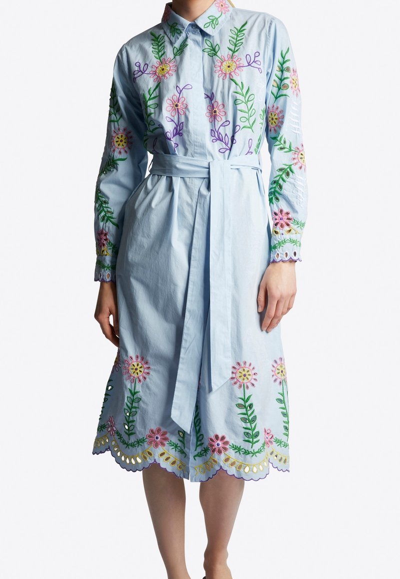 Abito camicia azzurro chiaro con maniche lunghe, ricami floreali in rosa e viola, e una cintura in vita. Presenta un orlo scallop e dettagli a taglio.