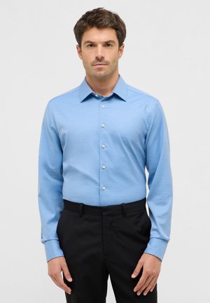 Hellblaues Langarmhemd aus glattem Stoff, mit einem klassischen Kragen und weißen Knöpfen, kombiniert mit schwarzen Hosen.