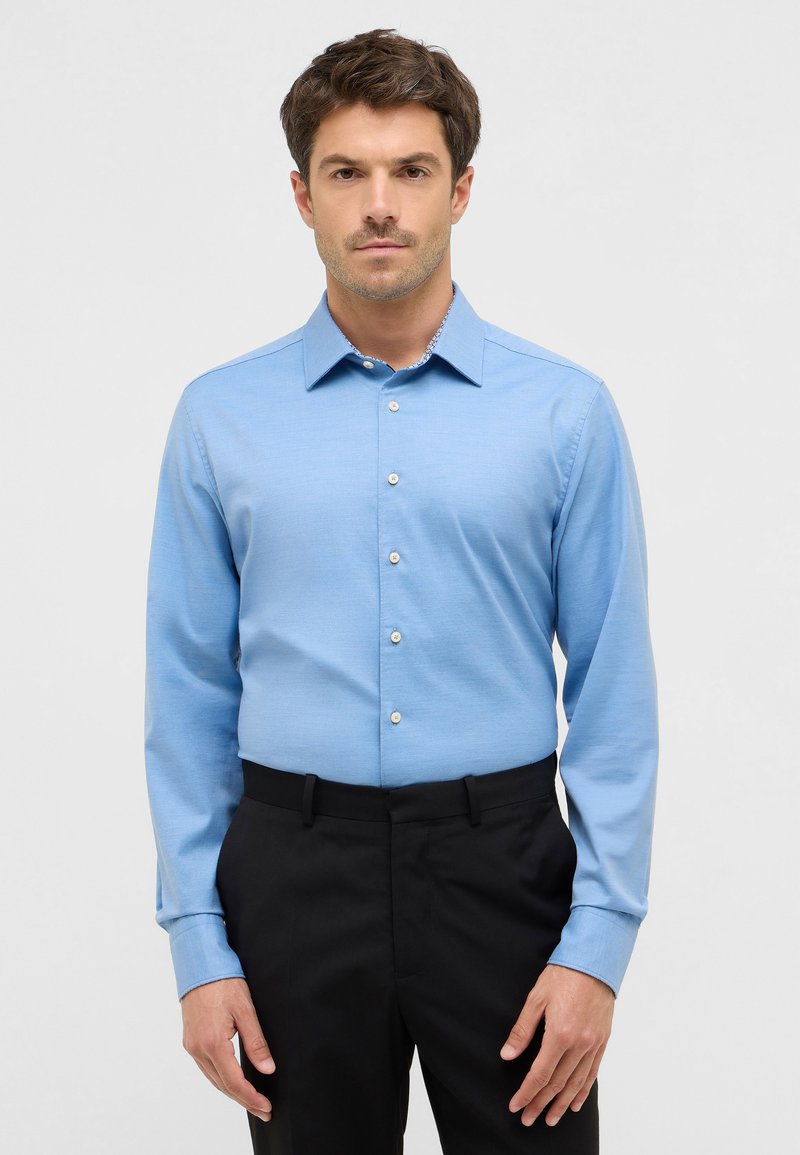 Camicia a maniche lunghe di colore azzurro chiaro, realizzata in tessuto morbido, con colletto button-down e bottoni bianchi, abbinata a pantaloni neri.