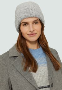 Grijze gebreide beanie met een textuurpatroon, gedragen met een grijze jas en een lichtblauw gestreept sweater. Kenmerkt zich door zachte stof en een comfortabele pasvorm.