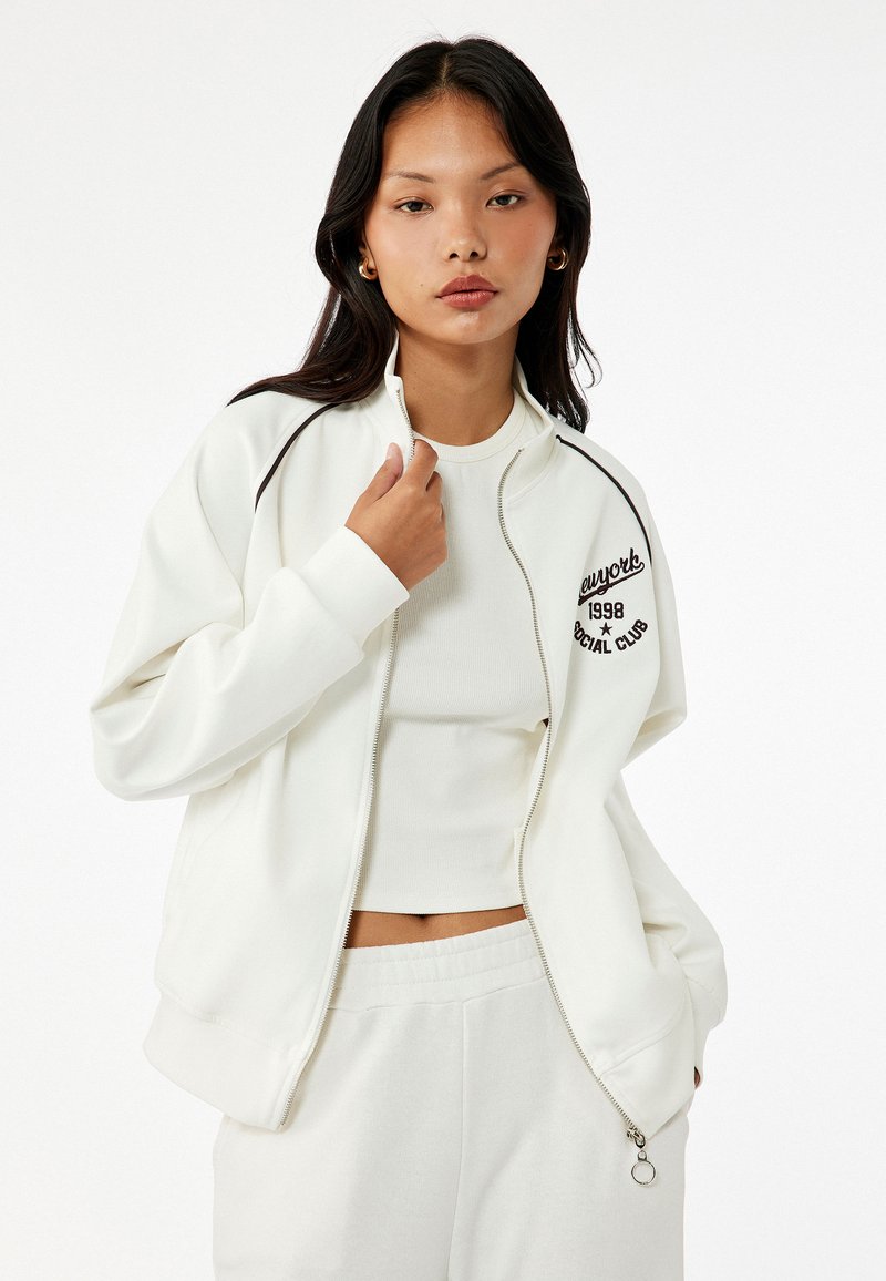 Weiße Zip-Jacke mit schwarzen Akzenten und gesticktem Logo auf der Brust. Kombiniert mit einem schmal geschnittenen weißen Oberteil und hellen Hosen.