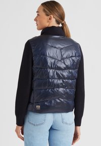 Frau mit hellbraunem Haar im Pferdeschwanz, die eine glänzende marineblaue Steppweste über einem schwarzen Pullover und hellblauen Jeans trägt, mit dem Rücken zur Kamera.