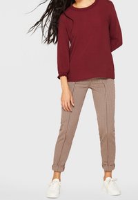 Burgundowy top z długim rękawem i okrągłym dekoltem, połączony z jasnobrązowymi wąskimi spodniami w paski i białymi trampkami. Gładkie tło.