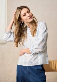 Femme aux longs cheveux blonds portant un chemisier à fleurs bleu clair et un jean bleu foncé, touchant ses cheveux, debout à l'intérieur contre un mur beige.