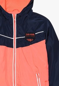 Námořnická a korálová větrovka se zipem vpředu, kapucí a bílými akcentními liniemi. Obsahuje vyšitý logopatch s "1982" na hrudi.