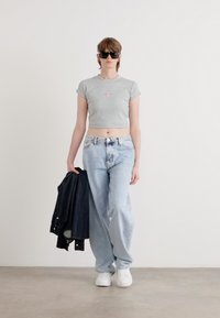 Siva kratki majica s malim ružičastim logom, lagane svijetloplave traperice, crna jakna i bijele tenisice. Model nosi crne sunčane naočale.