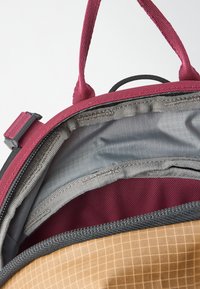 O interior da mochila apresenta um forro de tecido bordô, um compartimento cinza com fecho éclair e um fundo com padrão quadrado em cor bege. Alças texturizadas para melhor aderência.