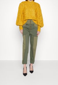 Geel gebreide trui met een gestructureerd patroon, gecombineerd met olijfgroene hoge taille broek met knoopdetails en een contrasterend paneelontwerp.