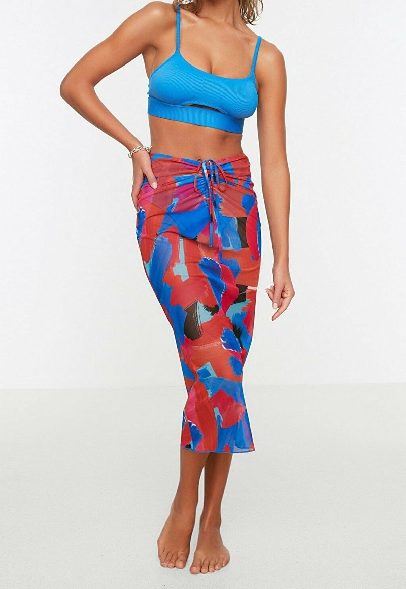 Femme portant un crop top bleu et une jupe portefeuille colorée à motif abstrait rouge, bleu et noir, debout pieds nus sur un fond uni.
