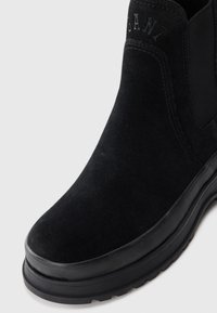 Schwarze Wildleder-Stiefelette mit Gummisohle, ausgestattet mit elastischen Seiteneinsätzen. Der Markenname ist am oberen Rand nahe dem Knöchel geprägt.