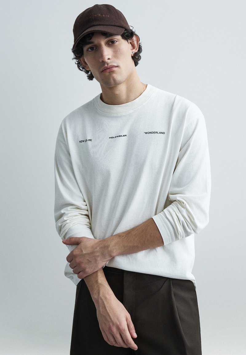 Camisola branca de algodão de manga comprida com detalhes em texto preto; boné castanho escuro. Modelo posa com os braços cruzados contra um fundo cinzento claro.