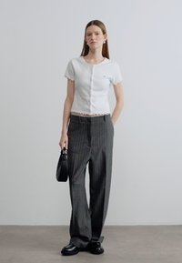 Λευκή μπλούζα με κοντά μανίκια και κυματιστές άκρες, με κλείσιμο με κουμπιά, συνδυασμένη με γκρι παντελόνια με ρίγες και φαρδιά πόδια και μαύρα παπούτσια.