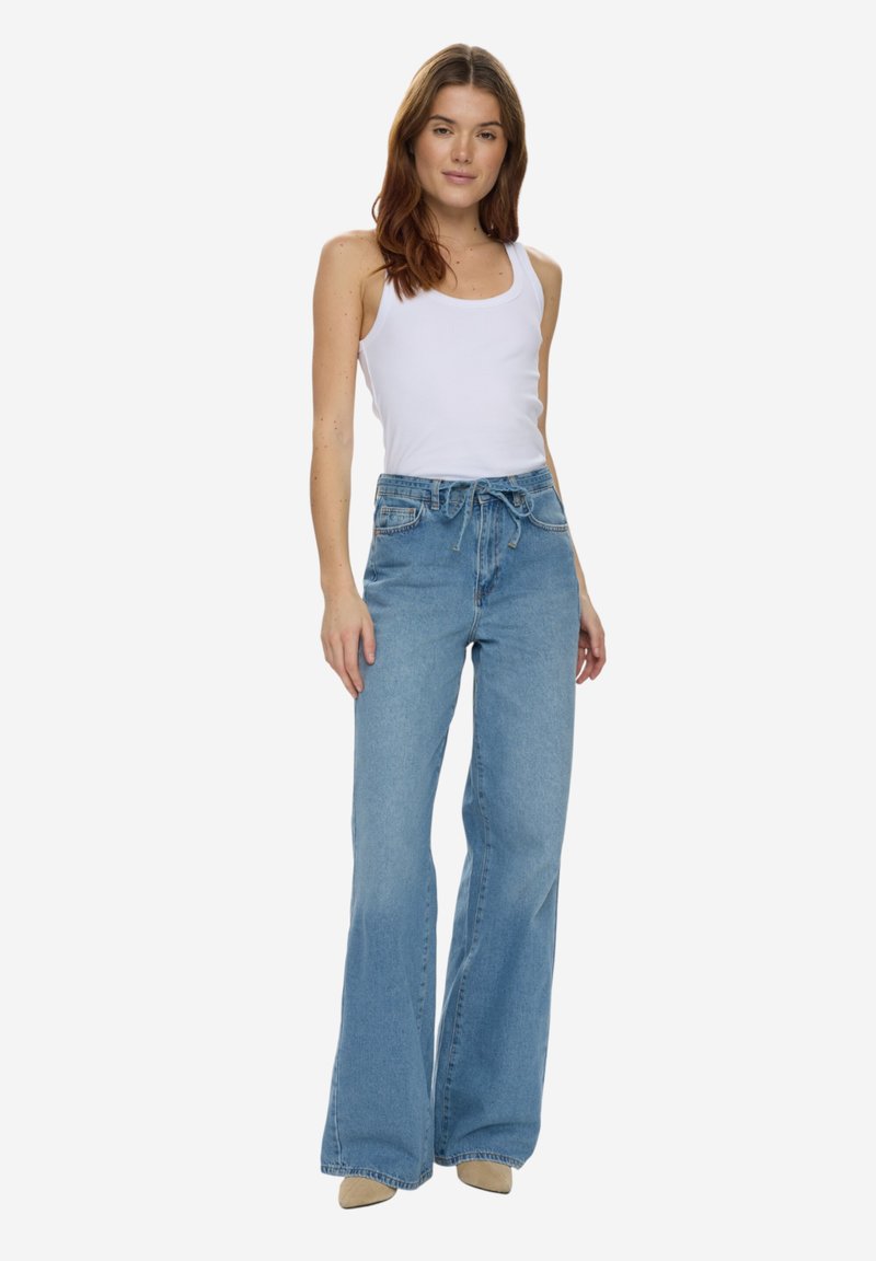 Femme debout portant un haut blanc sans manches, un jean bleu clair taille haute à jambes larges, et des chaussures pointues beiges sur un fond blanc.