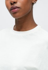 Λευκό T-shirt με στρογγυλή λαιμόκοψη, απαλή υφή, ορατές ραφές και άνετη εφαρμογή. Το μοντέλο φοράει ασημένιο κρίκο στο αυτί και έχει λεία χείλη.