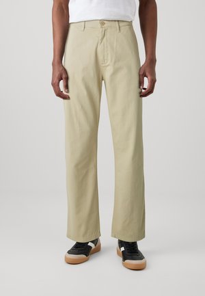 Lichtbeige, rechte broek met een gladde textuur en een enkele knoopsluiting. Gecombineerd met zwarte sneakers met een witte streep.