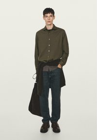 Camicia button-up verde oliva, canotta grigia, jeans blu scuro, scarpe marroni e un maglione marrone annodato in vita. Design semplice, vestibilità moderna.