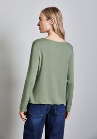Camicia a maniche lunghe in morbido tessuto verde chiaro con vestibilità rilassata, caratterizzata da un ampio scollo. Abbinata a pantaloni in denim blu scuro.