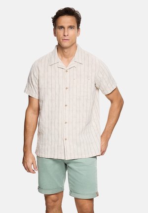 Hombre con camisa beige de manga corta abotonada con rayas sutiles y pantalones cortos verdes claros remangados, de pie contra un fondo blanco.